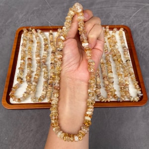 Peut inclure: Une main tient un collier de perles de citrine. Les perles sont un mélange de jaune clair et d'ambre, de forme irrégulière. D'autres rangées de perles similaires sont disposées sur un plateau en bois.