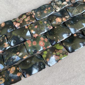 May include: A strand of rectangular beads made of dark green rhyolite stone, featuring natural inclusions in shades of reddish-brown, tan, and light green. Each bead has a smooth, polished surface with a glossy finish and rounded edges.