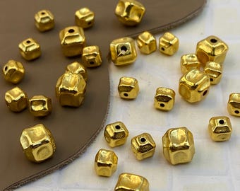 May include: A collection of small, metallic gold-colored beads with a cube-like, irregular shape. Each bead features a central hole for threading. The surface of the beads has a slightly textured, reflective finish with rounded edges and corners.
