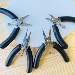 Puede incluir: Cuatro alicates de joyería con mangos negros y cabezas de metal plateado están dispuestos sobre una superficie blanca. Los alicates tienen diferentes formas de cabeza, adecuados para la fabricación de joyas y la artesanía.