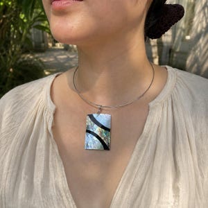Può includere: Una collana con pendente rettangolare caratterizzata da un intarsio in conchiglia di abalone iridescente con due strisce nere curve. Il pendente pende da un sottile girocollo in filo metallico color argento che riposa attorno al collo.
