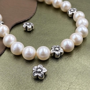 Può includere: Un braccialetto di perline composto da perle rotonde color bianco sporco alternate a piccoli distanziatori in metallo argentato a forma di fiore. Due perline in metallo argentato a forma di fiore sono posizionate vicino al braccialetto.