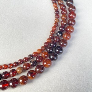 May include: Three strands of garnet gemstone beads in varying sizes. The beads range in color from deep red to reddish-brown, with a glossy finish. The strands are arranged in a curved pattern against a white background, showcasing the natural beauty of the stones.