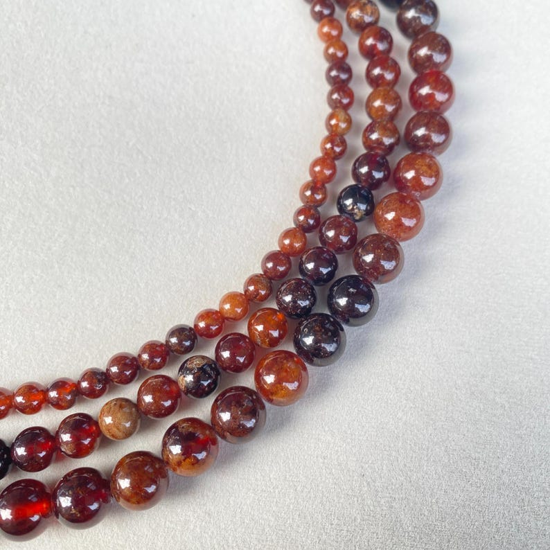 May include: Three strands of garnet gemstone beads in varying sizes. The beads range in color from deep red to reddish-brown, with a glossy finish. The strands are arranged in a curved pattern against a white background, showcasing the natural beauty of the stones.