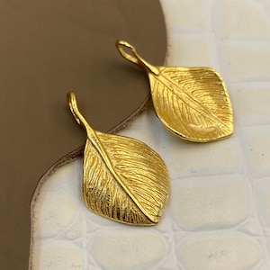 Puede incluir: Dos colgantes de metal en tono dorado con forma de hoja. Cada colgante presenta una superficie texturizada con una vena central y un pequeño lazo en la parte superior para su fijación. El diseño de la hoja tiene un acabado mate y forma orgánica.