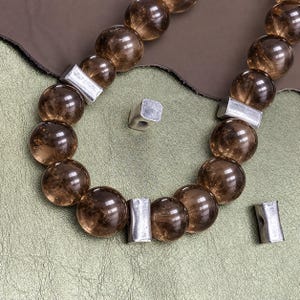 Può includere: Una collana composta da perline rotonde e traslucide di colore marrone, alternate a distanziatori rettangolari in metallo color argento. Le perline hanno una finitura liscia e lucida, mentre i componenti metallici presentano una texture opaca.