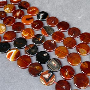 Peut inclure: Une collection de perles en pierre gemme plates et circulaires disposées en rangées. Les perles présentent une variété de couleurs, notamment le rouge profond, l'orange brûlé, le marron et le noir, avec des motifs d'agate naturelle et des centres cristallins.