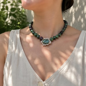 Peut inclure: Un collier de perles composé de pierres rondes à facettes vertes et rouge foncé. Le centre du collier présente une grande pierre ovale bleu-gris irisée, sertie dans une monture en métal argenté ornée de détails en forme de feuilles.