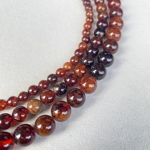May include: Strands of polished garnet beads in varying sizes and shades of red and brown. The beads range from a deep, dark red to a lighter, more translucent brown. The beads are strung together, creating a curved pattern against a white background.