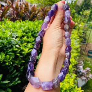 Può includere: Una collana di ametista sfaccettata con varie tonalità di viola. Le pietre preziose sono infilate insieme, creando un gioiello di grande effetto. Lo sfondo è un fogliame verde.