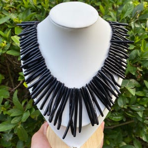 Può includere: Una collana importante caratterizzata da numerose perline nere cilindriche e allungate disposte a frangia. Le perline sono sospese da un punto centrale, creando un design denso e radiante che copre l'area del collo del supporto.