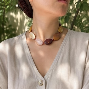 May include: A necklace featuring large, smooth, oval-shaped beads in shades of cream, pale pink, mustard yellow, and deep burgundy. The beads are separated by small, round, dark red spacer beads, creating a colorful and textured accessory.