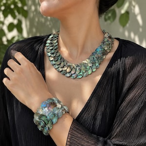 Può includere: Una collana e un bracciale a fascia larga abbinato, entrambi realizzati con dischi circolari sovrapposti di conchiglia di abalone. Le conchiglie presentano tonalità iridescenti di verde intenso, blu e viola, creando un aspetto testurizzato a scaglie.