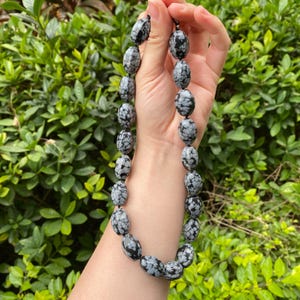 May include: A necklace of oval-shaped snowflake obsidian beads. The beads are primarily black with white and gray patterns, strung together on a dark cord. The necklace is held against a backdrop of green foliage.