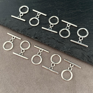 May include: A collection of nine silver-toned metal toggle clasps, each consisting of a circular ring and a matching T-bar component. These jewelry findings feature a smooth, polished finish and are arranged in a grid pattern for display.