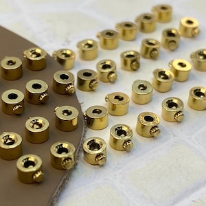 May include: A collection of small, cylindrical gold-toned metal crimp beads or cord ends, each featuring a tiny side screw mechanism. These metallic components are arranged in a grid-like pattern, showcasing their polished finish and circular shape.