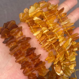 May include: Two strands of polished amber beads featuring irregular, chip-like shapes. One strand displays deep, dark brown tones, while the other strand showcases lighter, translucent golden-yellow hues. Both strands are strung on thin, flexible cord.