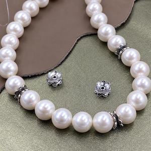 Può includere: Una collana composta da perle bianche rotonde alternate a copriperle in metallo argentato a forma di fiore. Due ulteriori copriperle in metallo argentato a forma di fiore sono posizionati sciolti al centro del filo di perle curvo.