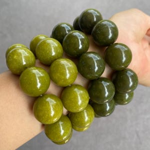 Può includere: Un bracciale a più fili composto da grandi perle sferiche e lisce in varie tonalità di verde oliva e verde bosco. Le perle hanno una finitura lucida e levigata e sono infilate insieme per creare un aspetto denso e stratificato.