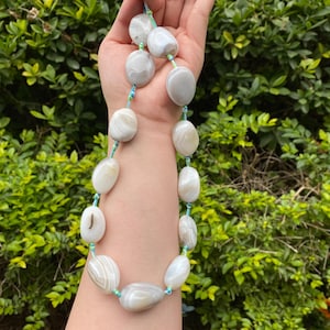 Könnte beinhalten: Eine Halskette mit großen, ovalen, grau-weißen Achatsteinen. Die Steine sind mit einer hellblauen und grünen Kordel aufgefädelt. Die Halskette wird vor einem Hintergrund aus grünem Laubwerk präsentiert.