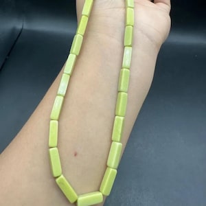 May include: A light green necklace made of rectangular beads. The beads are strung together. The necklace is shown on a person's arm against a dark background.