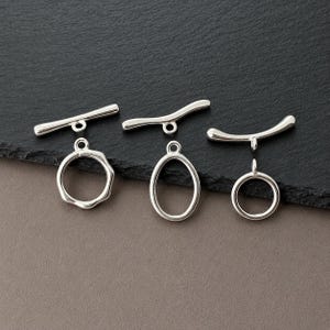 Può includere: Tre set di chiusure a T color argento per la creazione di gioielli. Ogni set è composto da una barra e un anello. Gli anelli variano nella forma, includendo un cerchio ondulato, un ovale liscio e un cerchio semplice, tutti con occhielli.