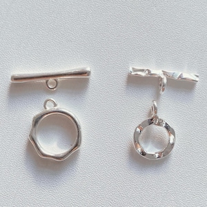 Puede incluir: Dos cierres de palanca plateados. Uno tiene una barra lisa y un anillo redondo con borde ondulado. El otro tiene una barra texturizada y un anillo redondo con borde ondulado. Ambos cierres están sobre una superficie blanca.