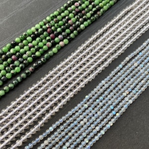 May include: Close-up of several strands of beads. One strand is green with red flecks, another is clear, and the last is a light blue. The beads are small and round, and the strands are arranged diagonally.
