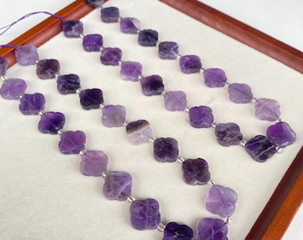 May include: Three strands of amethyst gemstone beads. Each bead is a square shape with rounded edges, strung on a purple cord. The beads are various shades of purple, from light lavender to deep violet. The beads are displayed on a white surface inside a wooden tray.
