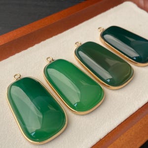 Könnte beinhalten: Vier grüne Jade-Anhänger mit goldfarbenen Rahmen. Jeder Anhänger ist rechteckig mit abgerundeten Kanten und einer kleinen Schlaufe oben. Der Jade-Farbton variiert von hell- bis dunkelgrün.