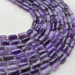 Puede incluir: Una colección de múltiples hilos de cuentas de amatista pulidas, cada una con una forma rectangular ligeramente irregular. Las cuentas muestran varios tonos de púrpura, desde lavanda claro hasta violeta intenso, ensartadas en un hilo púrpura.