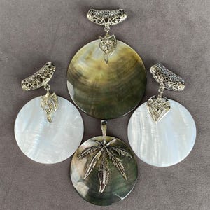 Op de afbeelding: Een sieradenset bestaande uit twee ronde witte parelmoeren hangers met sierlijke zilverkleurige filigraan oogjes, een grote ronde donkere iriserende hanger en een kleinere donkere iriserende hanger met een zilverkleurige bladbedel.