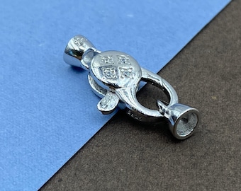 May include: A silver-toned lobster claw jewelry clasp featuring a circular center plate adorned with four small, clear, sparkling stones arranged in a square pattern. The clasp has a polished metallic finish and includes two conical end caps.