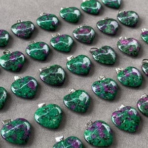 Puede incluir: Una colección de colgantes en forma de corazón hechos de piedra de rubí zoisita pulida. Cada colgante presenta una base verde vibrante con inclusiones moteadas de color magenta y negro, rematado con una pequeña anilla de metal plateado.