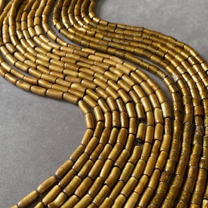 May include: Multiple strands of cylindrical beads in a metallic gold finish with a textured, speckled surface. The beads are arranged in a curved, flowing pattern, creating a dense, uniform appearance across the entire collection of strands.