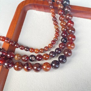 May include: Close-up of several strands of gemstone bead necklaces in varying sizes. The beads are a mix of deep red, brown, and amber hues, with a glossy finish. The necklaces are arranged on a wooden tray, showcasing their color variations.