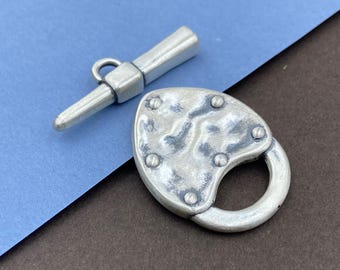 May include: A silver-toned toggle clasp set consisting of a T-bar component and a heart-shaped padlock component. The padlock features a textured, mottled surface with five small circular rivets arranged around the perimeter.
