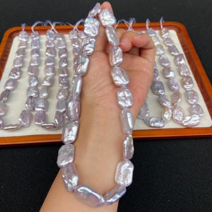 Può includere: Una collana di perle barocche iridescenti color lavanda è tenuta in mano. Diverse file di perle simili sono disposte su un vassoio di legno. Le perle hanno una forma oblunga irregolare e una superficie lucida.