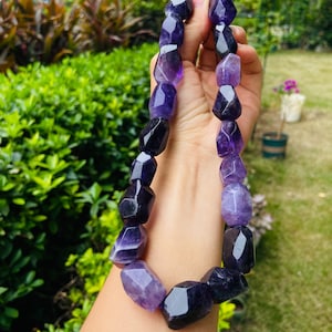 Puede incluir: Un collar compuesto por grandes cuentas de amatista facetadas e irregulares en tonos variados de morado intenso y lavanda claro. Las piedras están ensartadas en una sola hebra, mostrando variaciones naturales en color, claridad y forma.