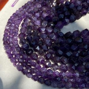 Könnte beinhalten: Nahaufnahme von Strängen facettierter Amethystperlen. Die Perlen sind tiefviolett und haben eine geometrische Form. Die Edelsteine sind aufgereiht und erzeugen einen geschichteten Effekt. Die Perlen sind in einem geschichteten Muster angeordnet.