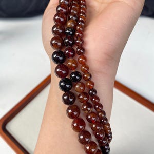 May include: Three strands of gemstone beads in varying sizes and shades of red-brown and black. The beads are round and appear to be strung together. The beads are held in a hand, with a wooden frame in the background.