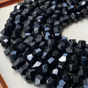 Peut inclure: Une collection de nombreuses petites perles noires polies avec une forme géométrique irrégulière à facettes. Chaque perle présente de multiples surfaces planes qui reflètent la lumière, créant un aspect brillant sur l'amas dense de pierres sombres.
