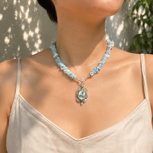 May include: A necklace featuring light blue, irregular stone beads strung together with a silver-toned toggle clasp. A central oval pendant hangs from the clasp, showcasing an iridescent abalone shell inlay set within an ornate silver-toned frame.