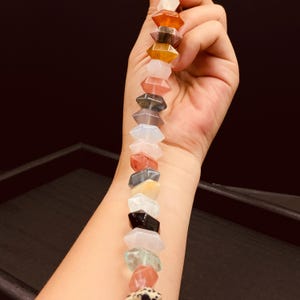 May include: A string of multi-coloured, faceted gemstone beads, including orange, yellow, pink, and grey. The beads are held up against a dark background, highlighting their unique shapes and natural variations. The gemstones are arranged in a vertical line.