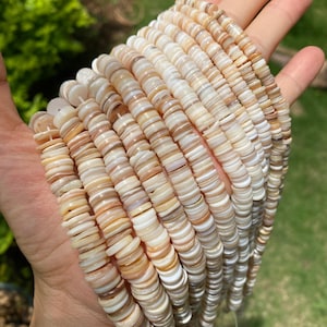 Pode incluir: Fios de contas de concha natural em tons de creme, bege e castanho. As contas planas em forma de disco são enfiadas, criando uma aparência texturizada. As contas são seguradas em uma mão.
