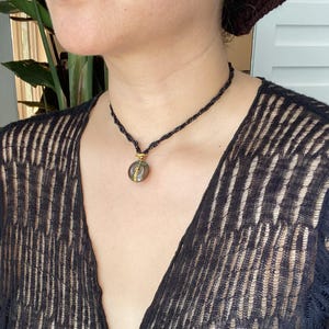 Op de afbeelding: Een zwarte kralenketting met een goudkleurige hanger. De hanger heeft een ronde, doorschijnende steen met een goudkleurig accent. De ketting wordt gedragen met een zwarte top met open weefsel.