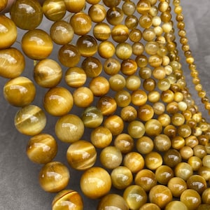 May include: A collection of spherical tiger's eye gemstone beads arranged in a gradient of sizes. The beads feature a polished, chatoyant surface with golden-brown bands and a reflective lustre, showcasing the natural fibrous structure of the stone.