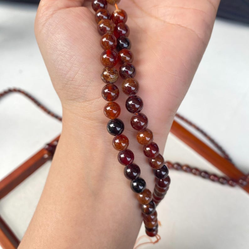May include: Strands of reddish-brown gemstone beads. The beads are round and vary in color from dark red to amber. The beads are strung together on a thin, light-colored string. The background is a light color.