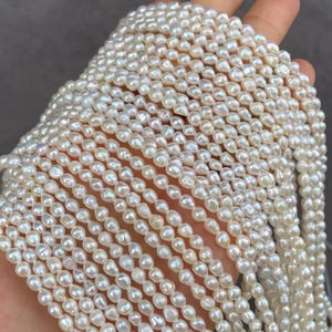 Pode incluir: Uma coleção de múltiplos fios de pequenas pérolas de água doce de cor esbranquiçada, com formas barrocas irregulares. Cada pérola individual apresenta uma superfície lustrosa e iridescente com uma textura ligeiramente irregular, disposta em filas paralelas.