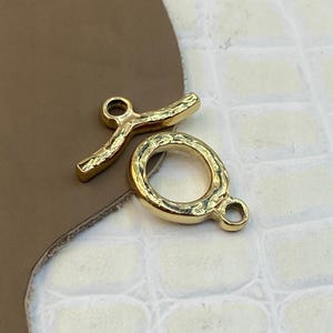 Puede incluir: Un conjunto de cierre de palanca de metal en tono dorado que consta de un componente de barra en T y un componente de anillo circular. Ambas piezas presentan una superficie texturizada y martillada. La barra tiene un bucle central y el anillo un ojal.